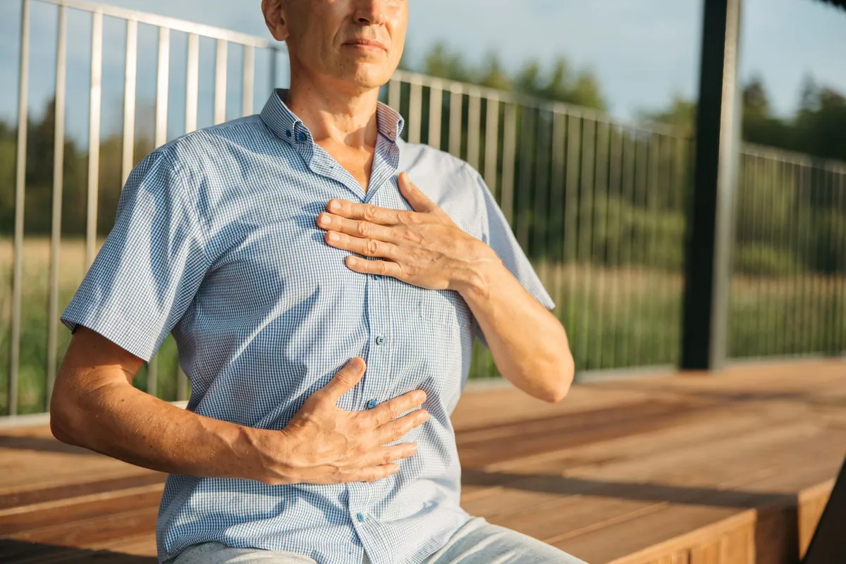 Older man sitting outdoors with one hand on his chest and one on his abdomen, practising a breathing technique to clear mucus